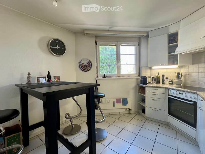 Modern kitchen with white cabinets, a black island table, and various appliances including an oven and a refrigerator. The space features tiled flooring and a window with a view of greenery.