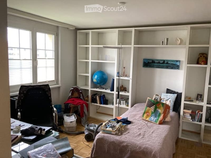 A furnished bedroom with a bed, a bookshelf, a chair, a globe, a table, a lamp, a mirror, and a picture frame. The room has wooden flooring and large windows with blinds.