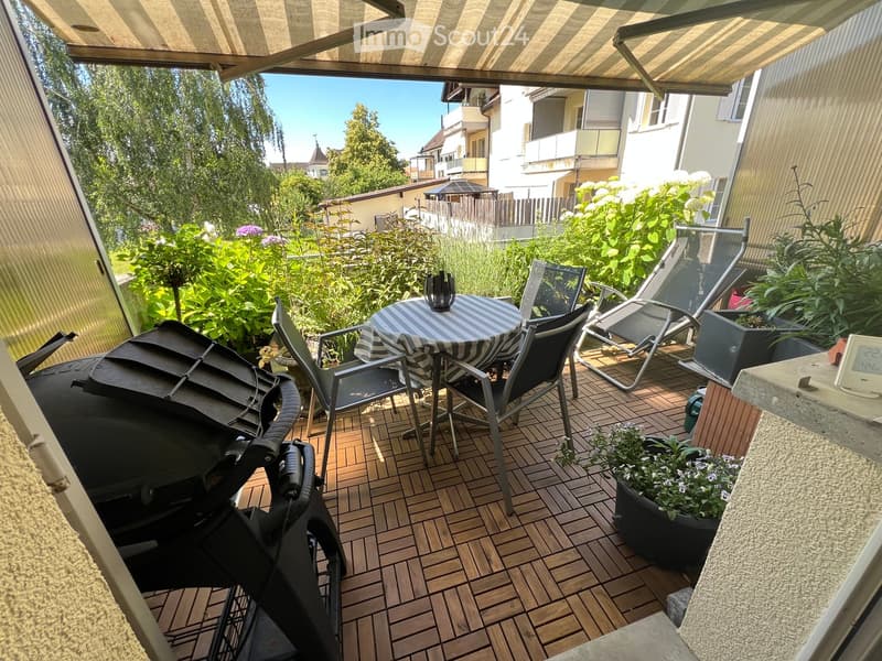 Balcony with wooden decking, round table with striped tablecloth, chairs, barbecue, and potted plants.