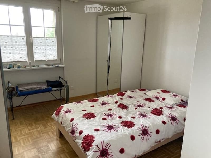 Bedroom with wooden flooring, a bed with red and white floral bedding, a wardrobe, a bench, and a mirror.
