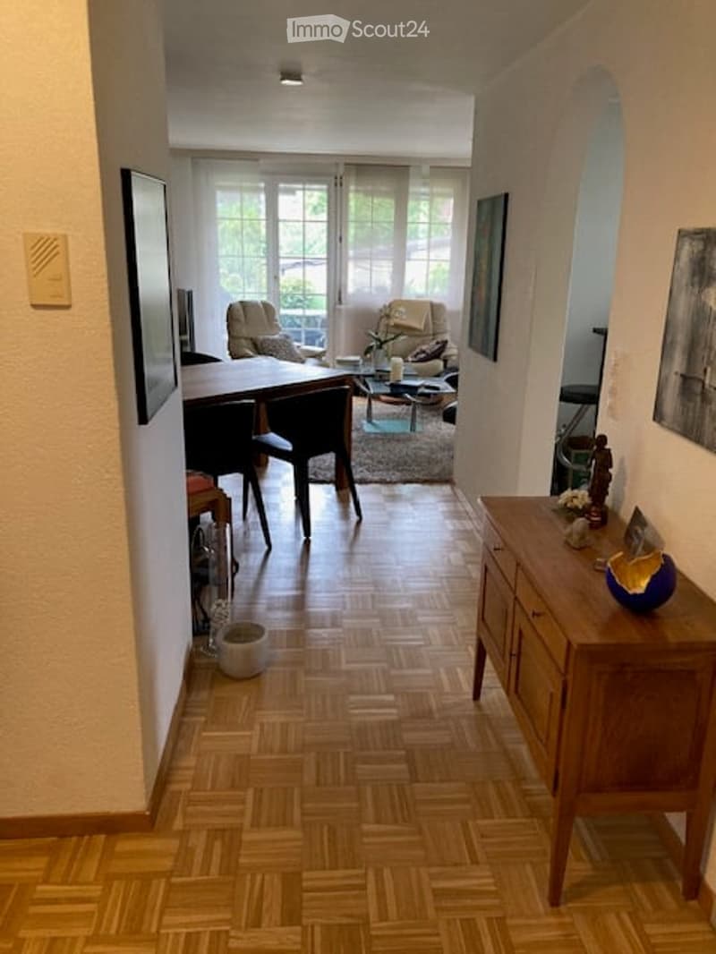 A well-lit hallway leading to a furnished room with a wooden floor, a large table, chairs, a sofa, a lamp, and a cabinet with a vase on top.