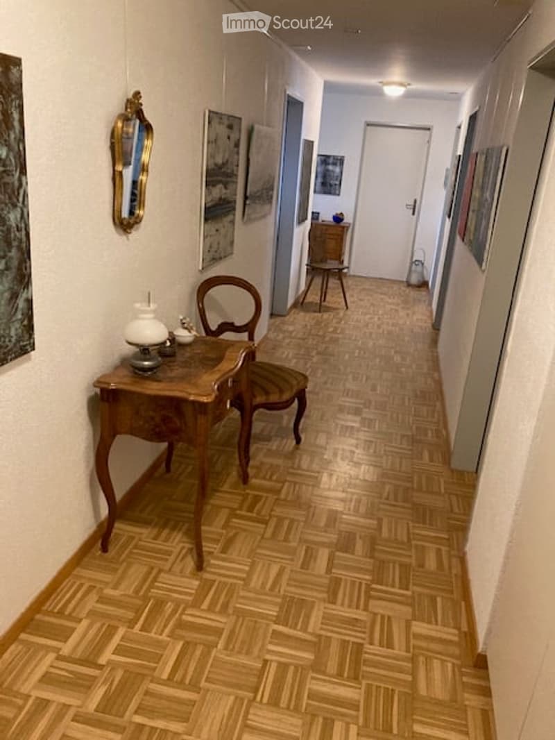 A narrow hallway with wooden parquet flooring, white walls, and several framed artworks. There is a small wooden desk with a lamp and decorative items, a chair, and a mirror on the wall.