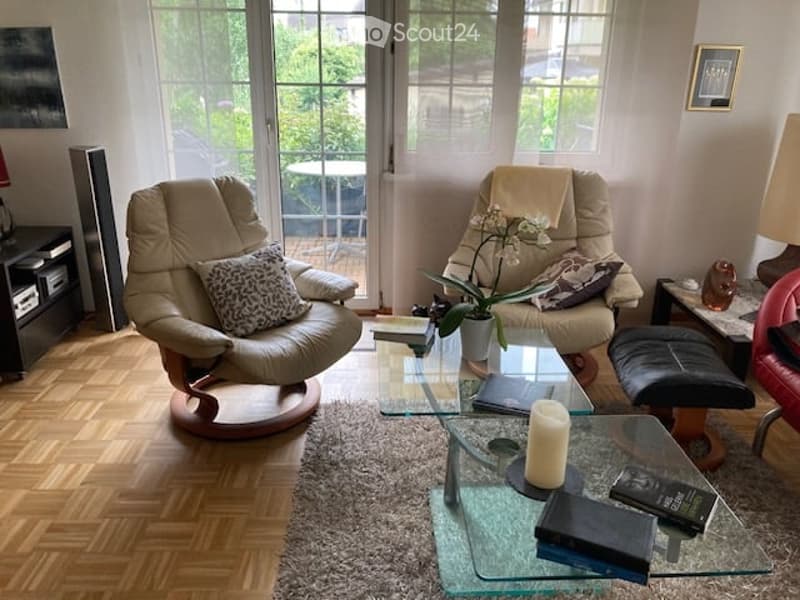 Modern living room with large windows and balcony access, featuring a beige leather sofa, glass coffee table, armchair, ottoman, and decorative items.