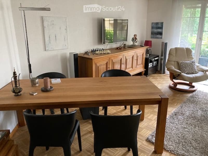 A modern, well-lit living room with a large wooden dining table, black chairs, a cream-colored sofa, a wooden cabinet, and a floor lamp. The room features large windows and a cozy, inviting atmosphere.