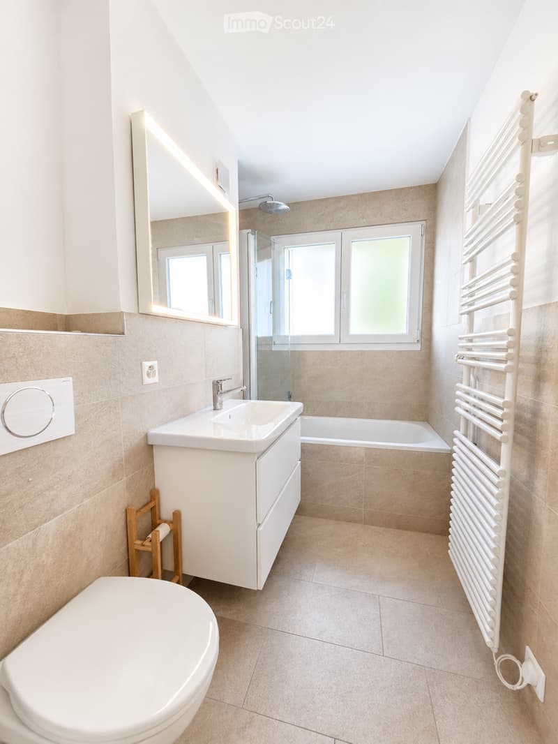 Modern bathroom with beige tiles, white fixtures, a large mirror, a bathtub, a toilet, a sink, and a radiator.