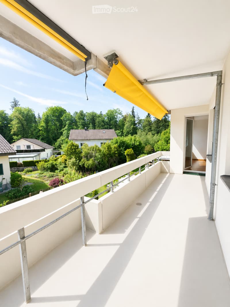 Bright balcony with white flooring, metal railing, and yellow retractable awning, overlooking a green garden and neighboring houses.