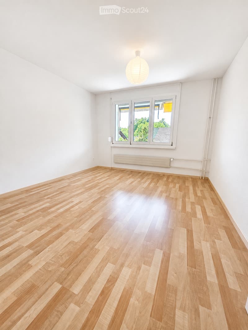 Empty room with light wooden flooring, white walls, and large windows letting in natural light. A single pendant light fixture hangs from the ceiling.