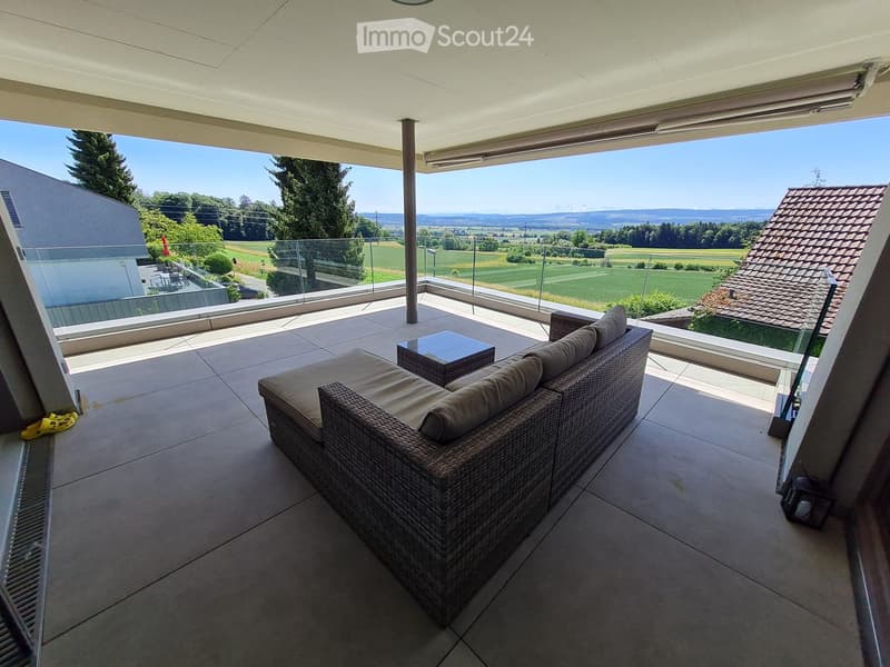 Balcony with a wicker sofa, a small table, and a scenic view of green fields and hills.