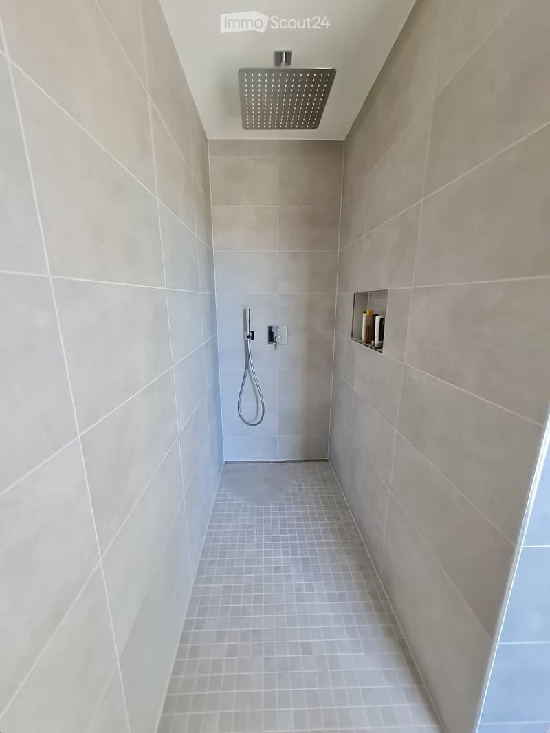 Modern shower area with grey tiled walls and floor, featuring a large overhead showerhead and handheld shower hose.