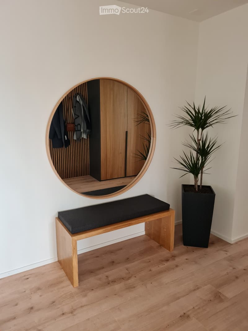 A modern hallway with a wooden bench, a round mirror, and a potted plant. The floor is made of light wood, and the walls are white.