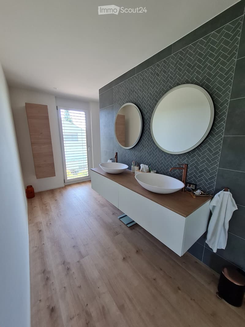 Modern bathroom with two sinks, a wooden countertop, white cabinets, and two large round mirrors on a dark tiled wall. The floor is light wood, and there is a towel hanging on the side.