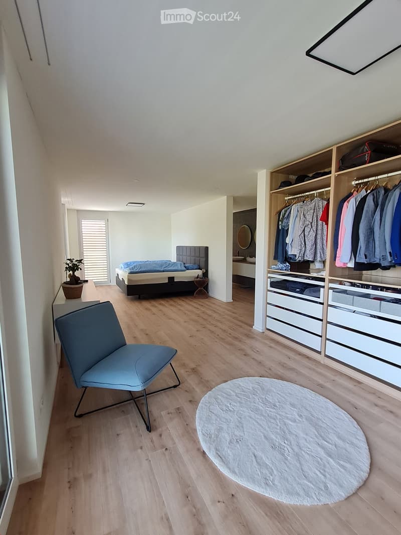 Modern bedroom with light wooden flooring, a bed with a dark headboard, a blue armchair, a round white rug, a built-in wardrobe with hanging clothes and drawers, and a small plant on a side table.