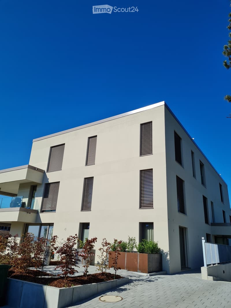 Modern two-story house with white exterior, large windows, and a balcony on the upper level. The house features a clean, minimalist design with a raised ground floor and a small landscaped garden area in front.