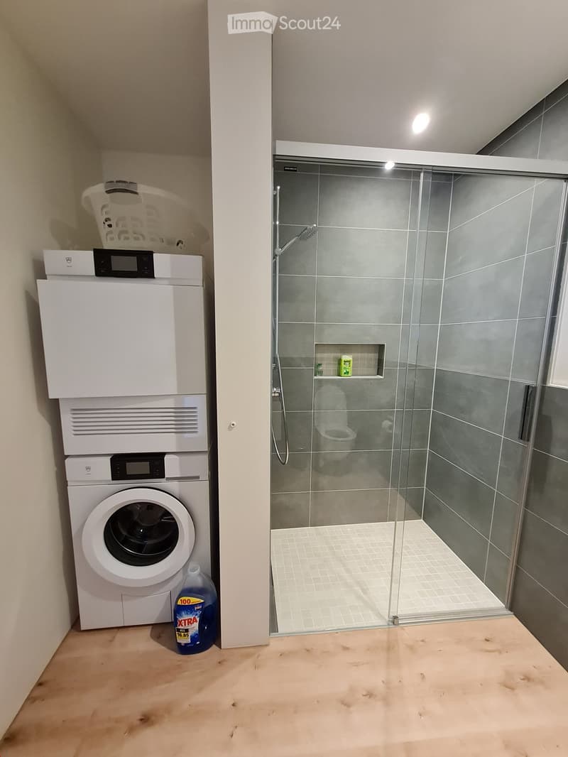 Modern bathroom with a glass-enclosed shower, toilet, and a stacked washing machine and dryer unit. A cleaning bottle is placed on the floor next to the washing machine.