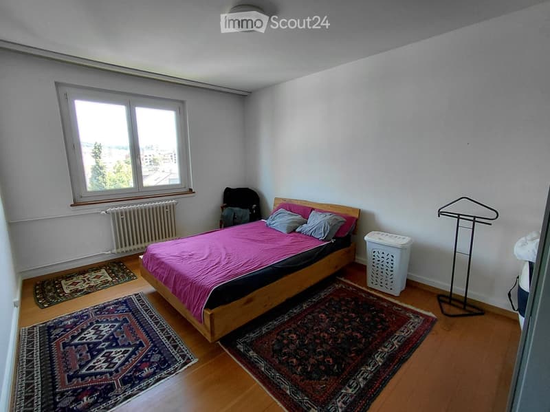 Bedroom with wooden floor, bed with pink bedding, pillows, a chair, radiator, coat rack, laundry basket, and rugs.