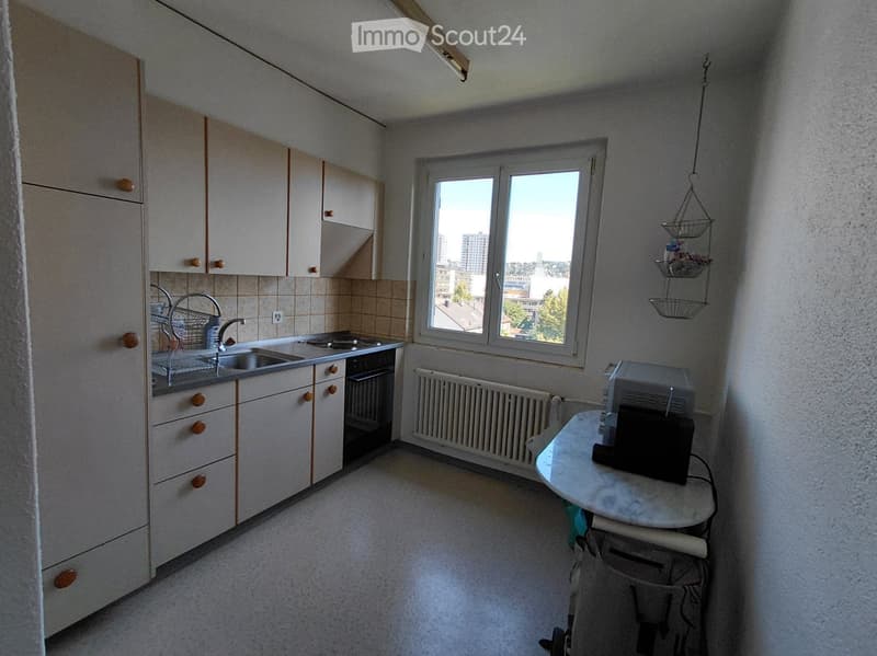 A small kitchen with white cabinets, a sink, an oven, a table with a microwave, and a window overlooking a cityscape.