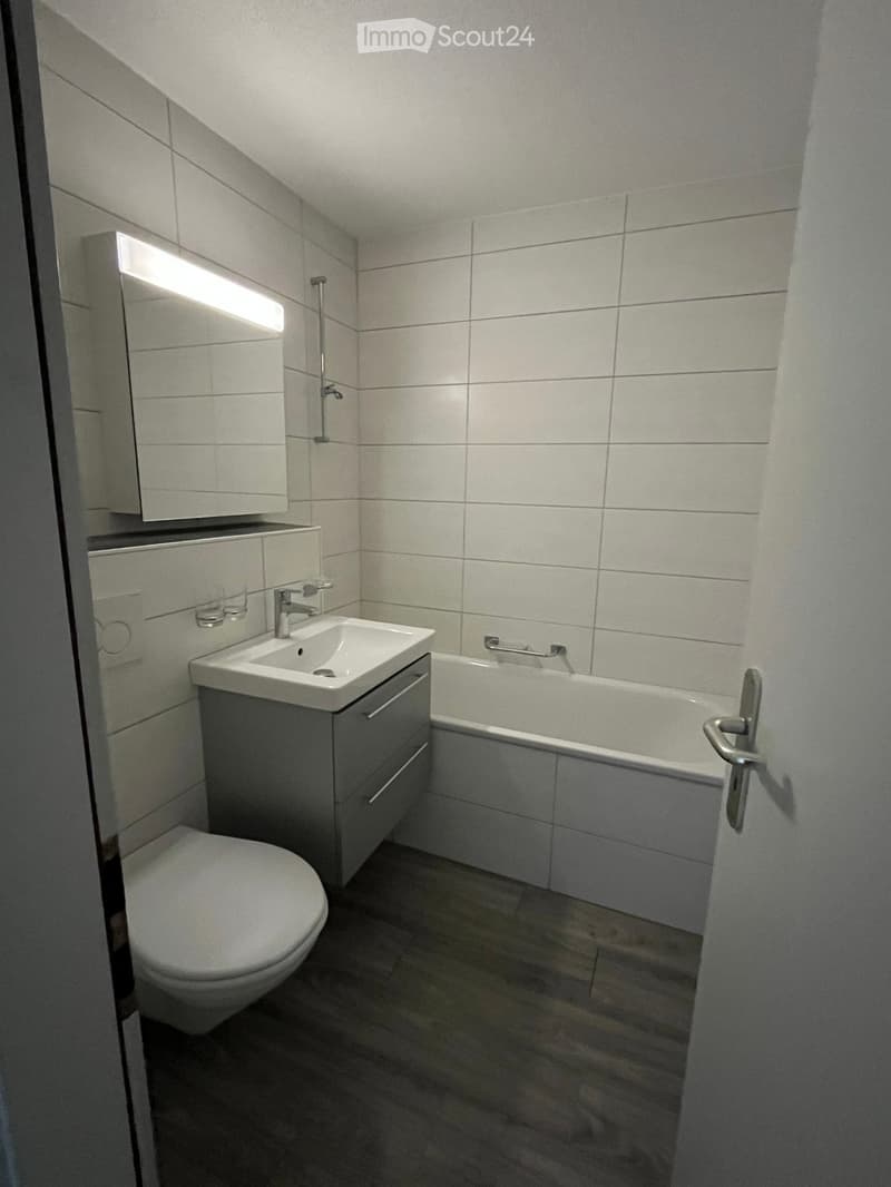 Modern bathroom with white tiles, a toilet, a sink with a cabinet, a bathtub, and a mirror with built-in lighting.