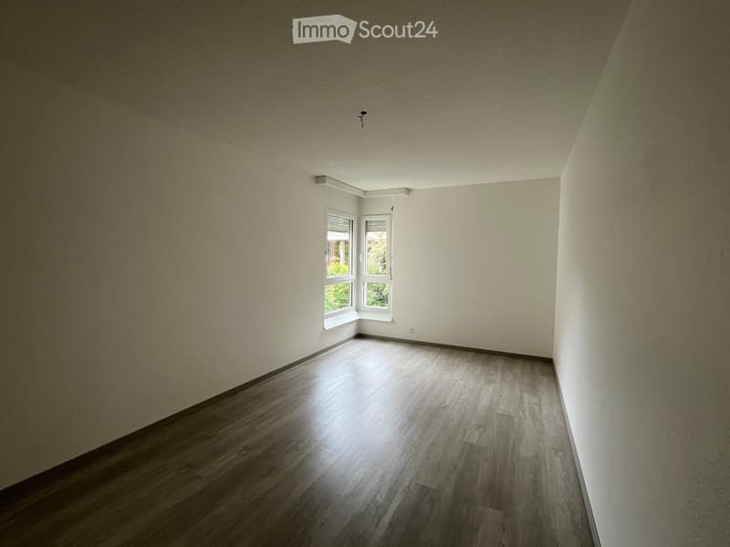 Empty room with light-colored walls and wooden flooring, featuring a window with blinds and a small light fixture on the ceiling.