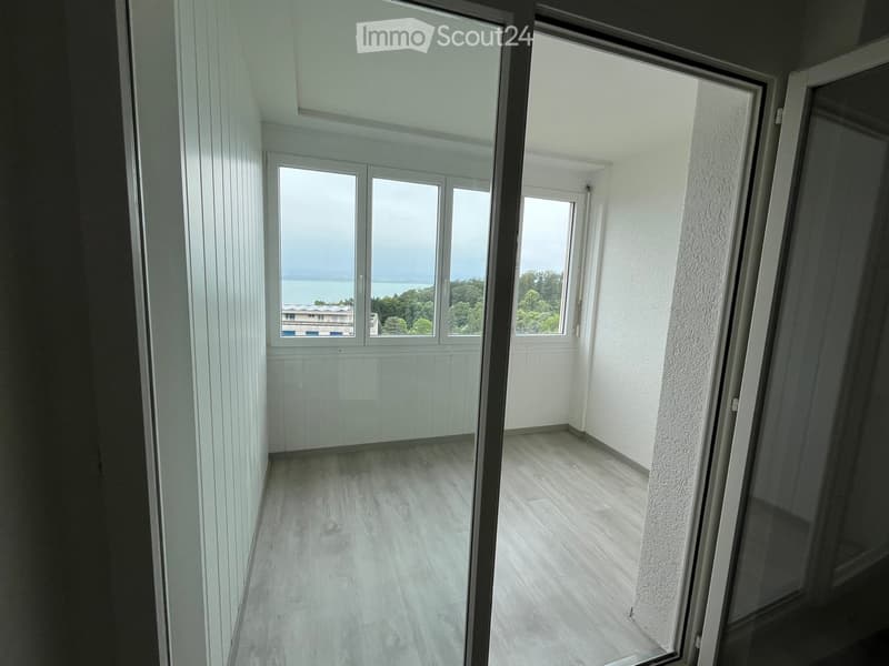 Empty unfurnished room with light wooden flooring, white walls, large windows, and a view of a lake and trees.