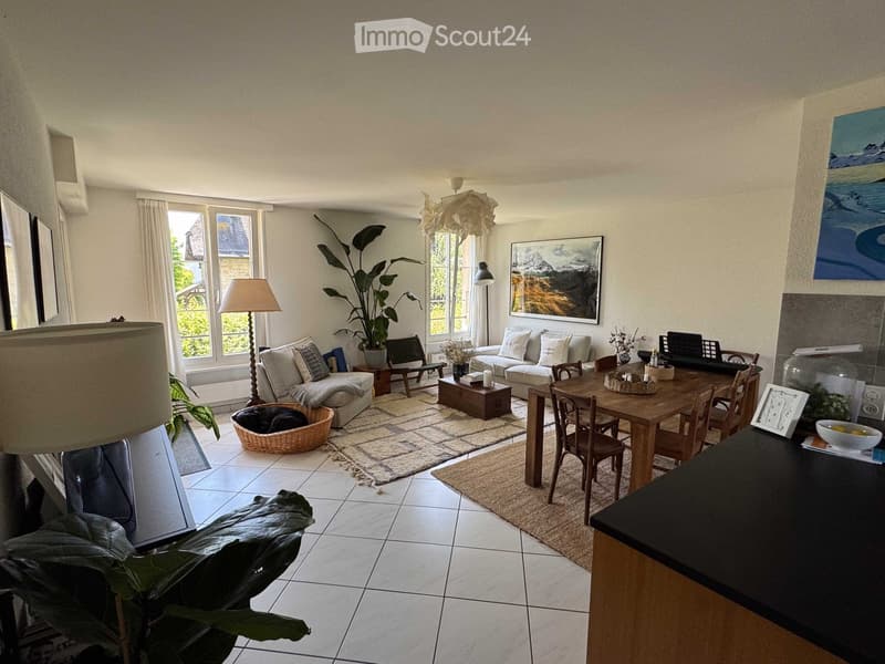 Bright living room with large windows, white walls, and a mix of modern and rustic furniture including a sofa, coffee table, dining table, and chairs.