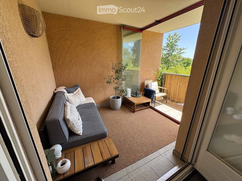 Balcony with a grey sofa, wooden table, small plant, and a view of greenery.