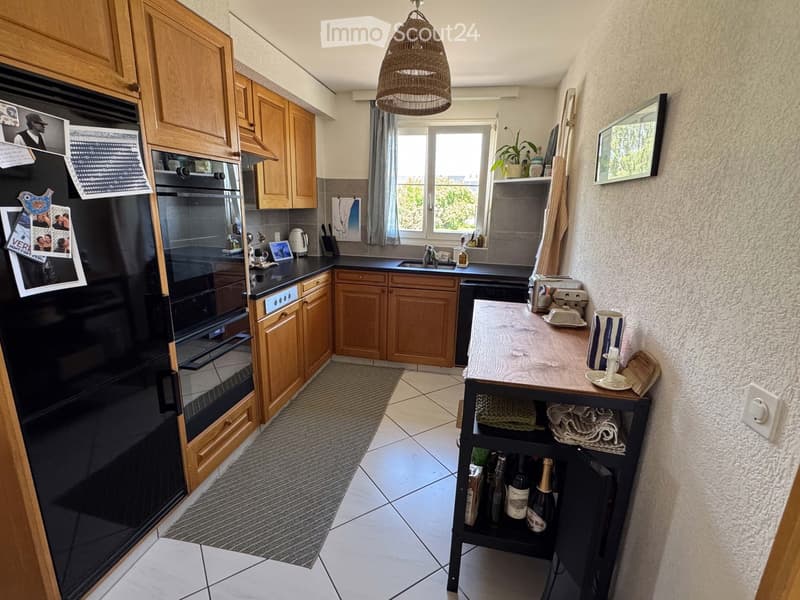 A modern kitchen with wooden cabinets, stainless steel appliances, a central island, and a window with a view. The space includes a refrigerator, oven, sink, and various kitchen items.
