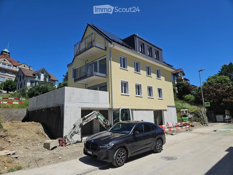 A multi-story residential building with a yellow facade, multiple windows, and balconies. A car is parked in front, and construction work is visible with an excavator and a partially constructed garage.