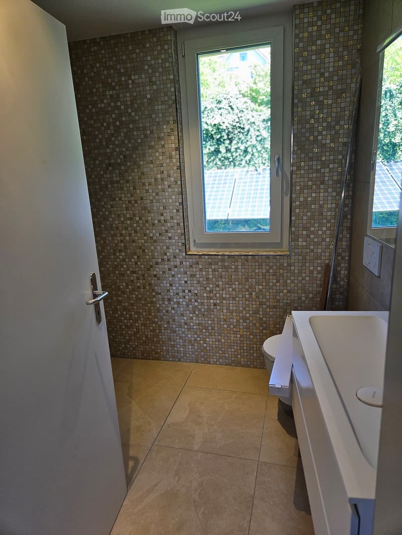 Modern bathroom with beige tiles, a white sink, and a toilet. Features a large window with a view of greenery and a tiled shower area.