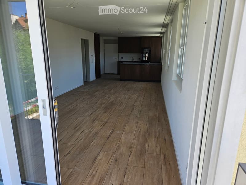 Empty hallway with wooden flooring, white walls, and a view into a kitchen area with dark cabinets and a sink.