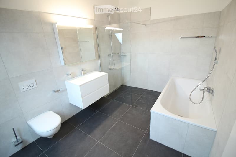 Modern bathroom with a white toilet, bathtub, sink, mirror, and glass-enclosed shower. Dark tiled floor and light tiled walls.
