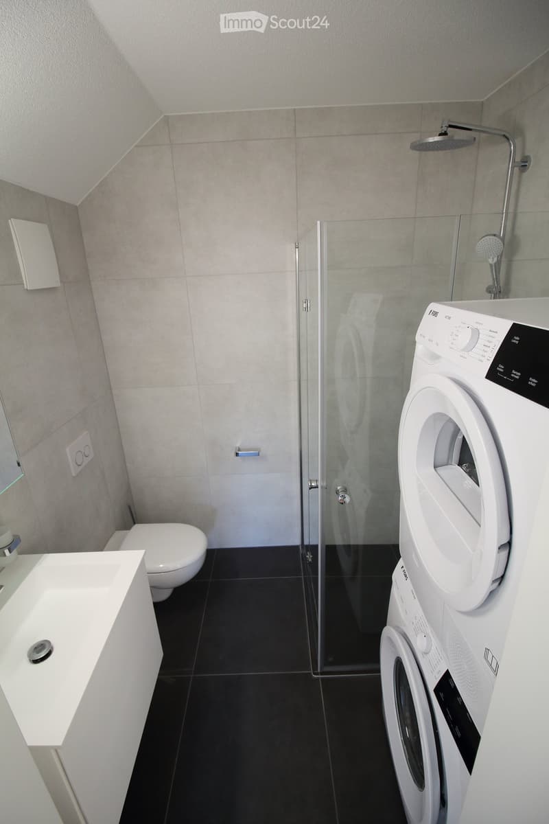 Modern bathroom with a toilet, sink, mirror, glass-enclosed shower, and a stacked washing machine and dryer unit.