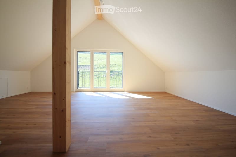 Empty attic room with wooden flooring, white walls, and large windows offering a view of the outdoors.
