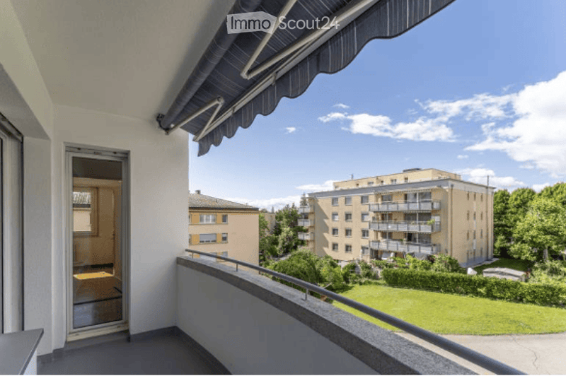 Balcony with a view of a residential area, featuring a metal railing, a sliding door leading to an interior space, and a clear blue sky with scattered clouds.