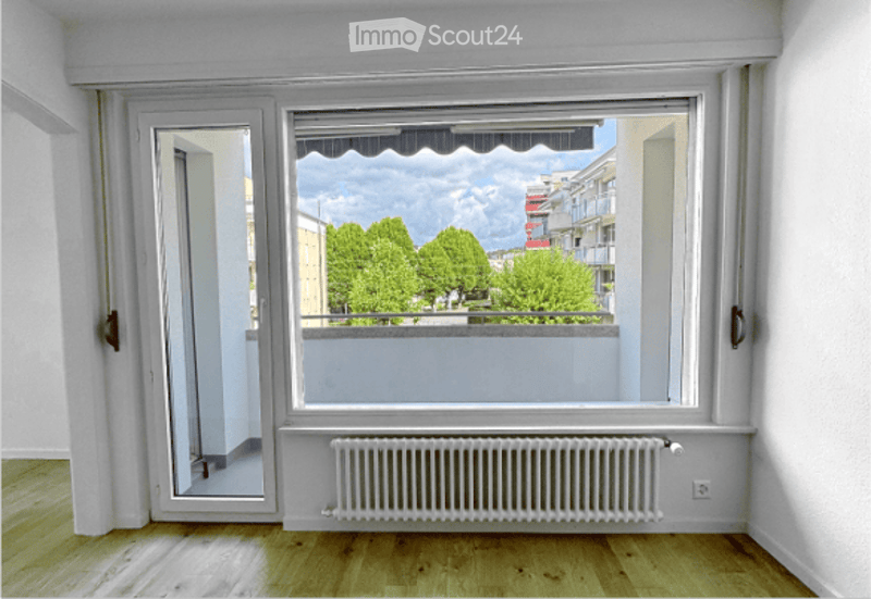 Empty room with wooden flooring, white walls, large window with balcony access, radiator, and a sliding door leading to the balcony.