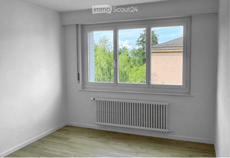 Empty room with light wooden flooring, white walls, and a large window with a view of greenery. A radiator is mounted on the wall below the window.