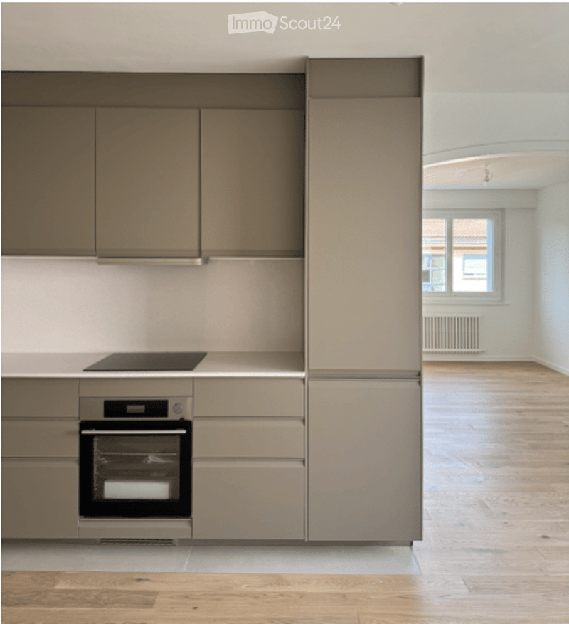 Modern kitchen with light grey cabinets, stainless steel oven, and refrigerator. The space features light wooden flooring and a neutral color palette.