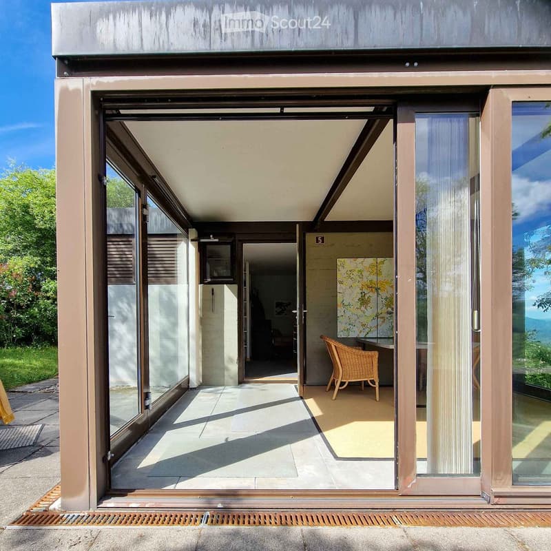 Modern glass-enclosed balcony with a wicker chair, large windows, and a view of greenery.
