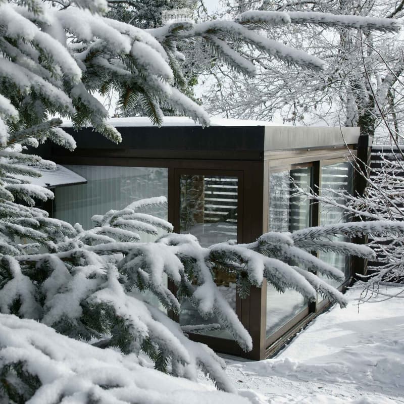 A modern, snow-covered building with large glass windows and a dark wooden frame, surrounded by snow-laden evergreen trees.