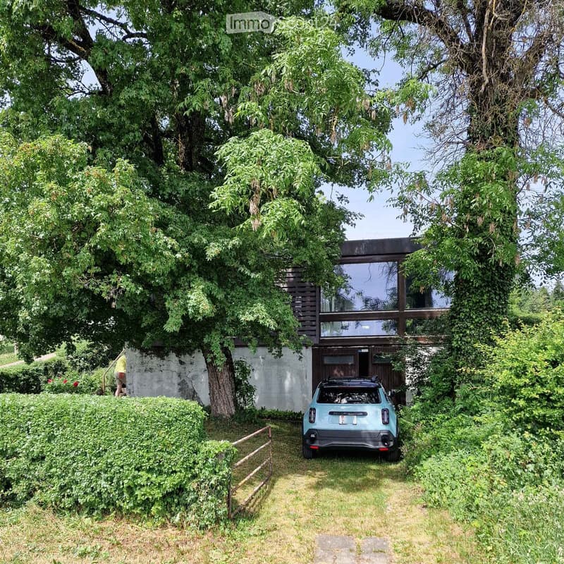 A modern two-story house with large windows, surrounded by lush greenery and trees. A light blue car is parked in front of the house.