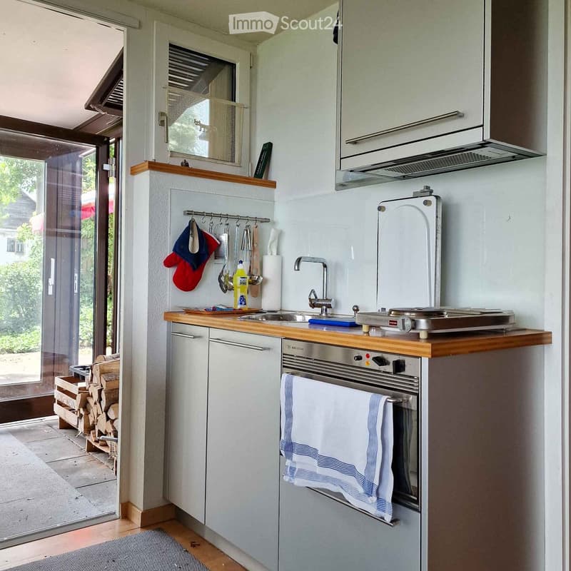 Compact kitchen with wooden countertops, stainless steel sink, oven, refrigerator, and hanging oven mitts. A towel is draped over the oven handle, and a bottle of cleaning solution sits on the counter.