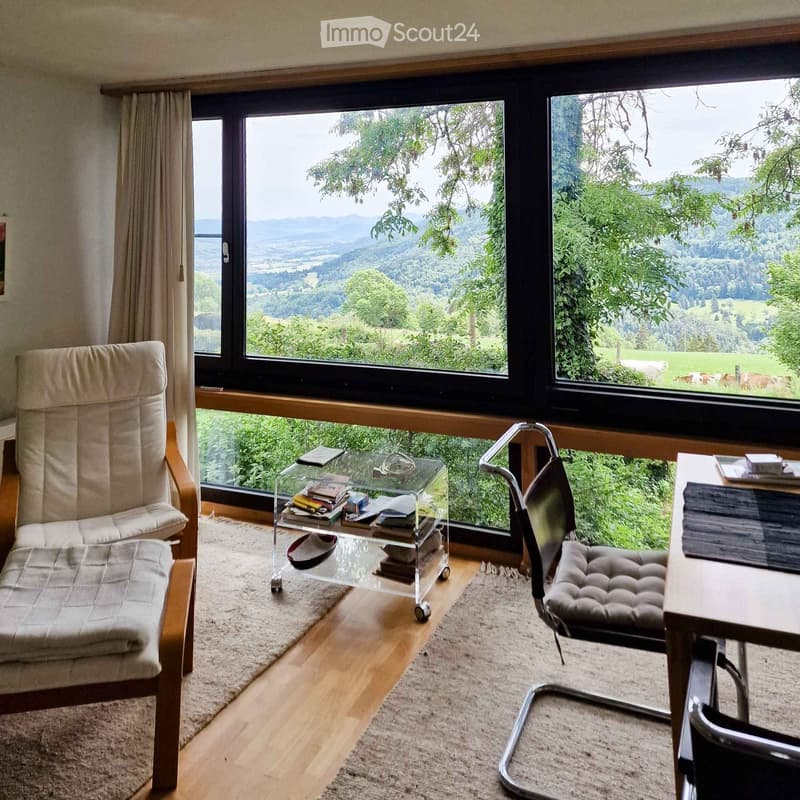 Bright room with large windows offering scenic views, furnished with a comfortable chair, a glass coffee table with books, a modern desk, and a sleek chair.