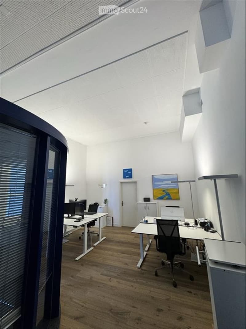Modern office space with wooden flooring, white walls, and large windows. Includes desks, chairs, computers, a floor lamp, and a painting on the wall.