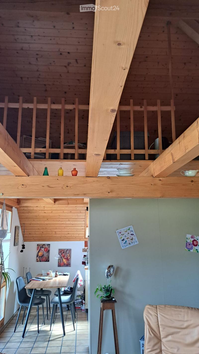 Open-plan living and dining area with wooden beams, a raised loft area, a dining table with chairs, a sofa, and decorative items on shelves.