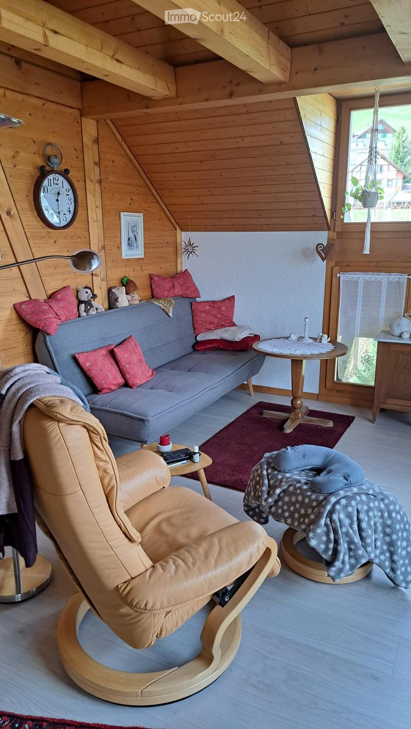 Cozy living room with wooden walls and ceiling, featuring a grey sofa with red cushions, a leather armchair, a small round table, and a clock on the wall.