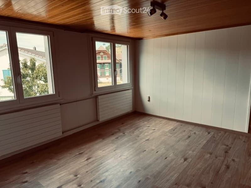 Empty room with wooden flooring, white walls, large windows, and a wooden ceiling.