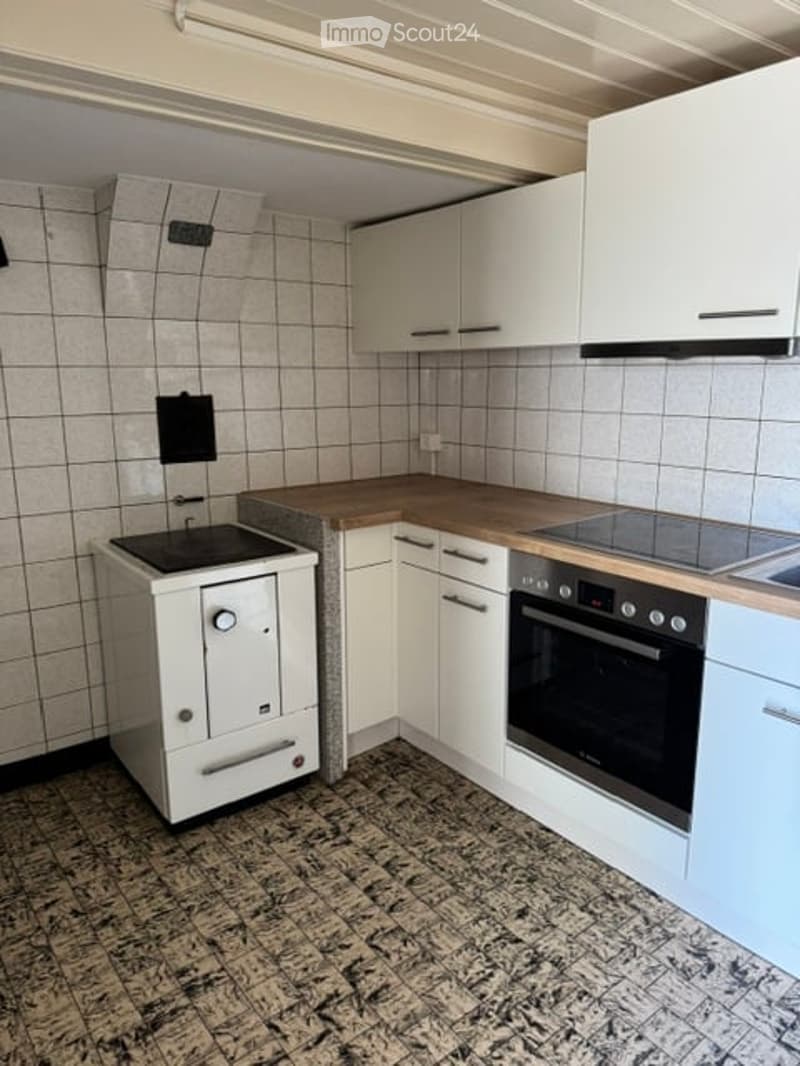 A compact kitchen with white cabinets, a wooden countertop, and built-in appliances including a stove and oven. The floor is tiled in a dark, patterned design, and the walls are covered with light grey tiles.