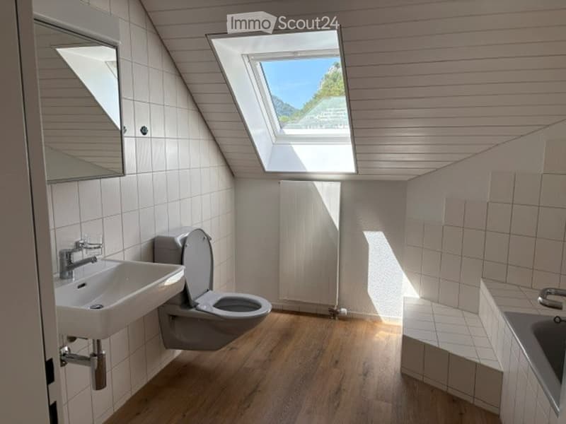Bright bathroom with wooden flooring, white tiled walls, a sink, toilet, and a skylight offering a scenic view.