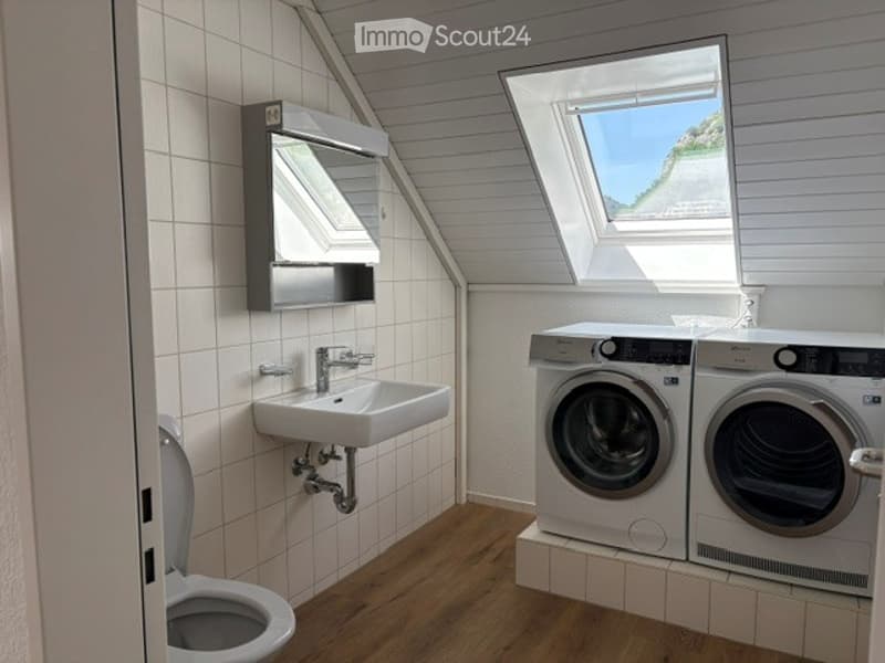 Bright bathroom with white tiled walls, wooden floor, a sink with a modern faucet, a toilet, a mirror cabinet, and a washer/dryer unit. Skylight windows provide natural light.