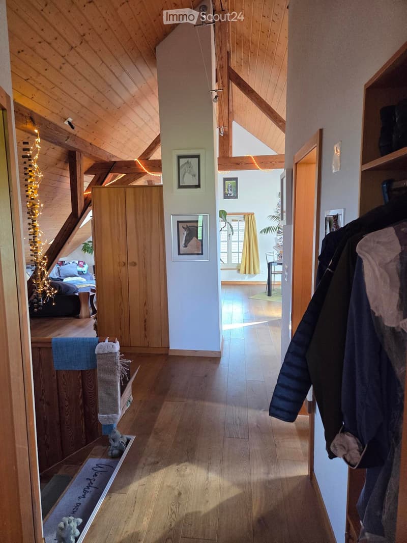 Bright hallway with wooden floors and exposed wooden beams, leading to a bedroom area with a bed and a living area with a chair and table.