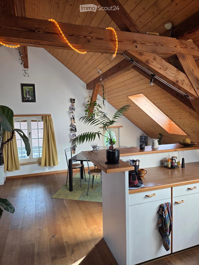 Bright, airy kitchen with wooden beams and slanted ceiling, featuring a central island with a wooden countertop, bar stools, and various kitchen items. There is a dining area with a table and chairs, and large windows with curtains letting in natural ligh